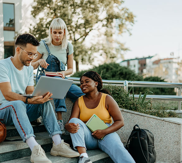 Mehrere Studierende sitzen gemeinsam im Freien und arbeiten an einem Laptop sowie an Lernunterlagen.