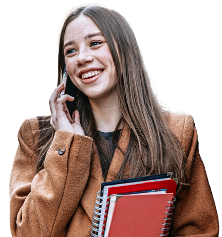 Eine junge Frau steht im Freien, telefoniert mit dem Smartphone und h&auml;lt mehrere Notizb&uuml;cher im Arm. Sie tr&auml;gt einen braunen Mantel und einen Rucksack und wirkt freundlich und entspannt.