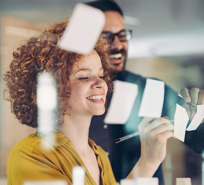 Zwei Personen stehen vor einer Glaswand und ordnen bunte Haftnotizen. Eine Person schreibt auf einen Zettel, w&auml;hrend die andere im Hintergrund l&auml;chelt. Die Szene vermittelt Teamarbeit und kreatives Brainstorming.