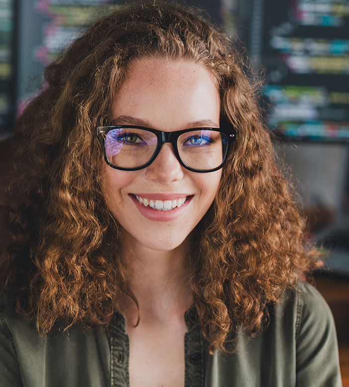 Eine Frau mit lockigen Haaren und Brille l&auml;chelt in die Kamera. Sie sitzt an einem Schreibtisch, im Hintergrund sind mehrere Bildschirme mit Programmcode zu sehen.