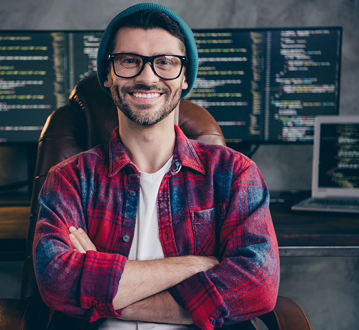 Ein Mann mit Brille und M&uuml;tze sitzt vor mehreren Monitoren mit Programmcode. Er l&auml;chelt und hat die Arme verschr&auml;nkt. Der Arbeitsplatz wirkt modern und technisch ausgestattet.
