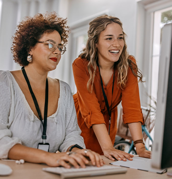 Zwei Frauen stehen an einem Schreibtisch in einem hellen B&uuml;ro und arbeiten gemeinsam an einem Computer. Beide tragen Namensausweise und wirken konzentriert und engagiert.