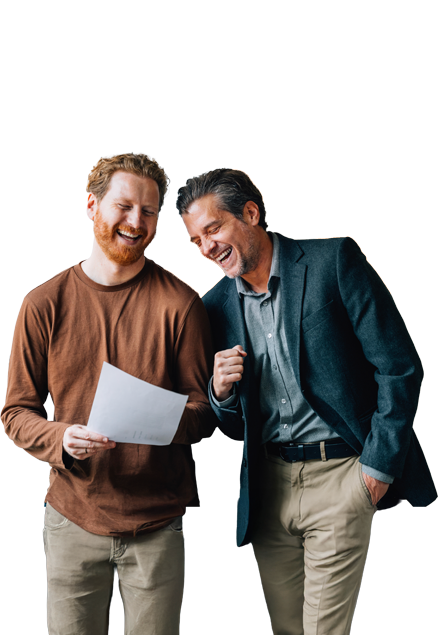 Zwei M&auml;nner stehen nebeneinander, lachen und betrachten gemeinsam ein Blatt Papier. Einer h&auml;lt das Dokument in der Hand, der andere zeigt gestikulierend darauf.