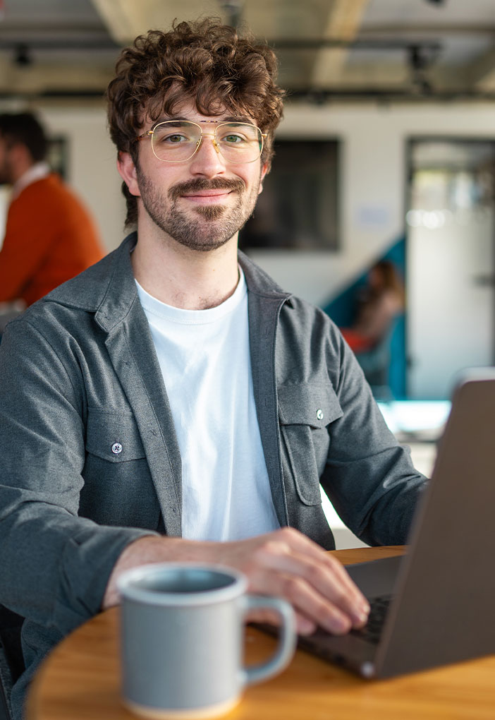 Ein Mann sitzt an einem Tisch in einem modernen Arbeitsumfeld und arbeitet an einem Laptop. Vor ihm steht eine Kaffeetasse, im Hintergrund sind unscharf weitere Personen zu sehen.