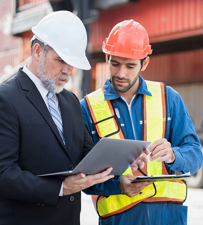 Zwei M&auml;nner mit Schutzhelmen stehen auf einem Betriebsgel&auml;nde mit Containern im Hintergrund. Einer h&auml;lt ein Tablet, der andere ein Klemmbrett. Beide besprechen gemeinsam Arbeitsunterlagen.
