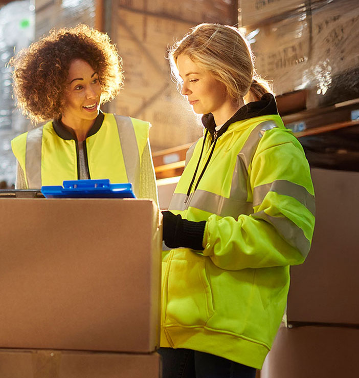 Zwei Frauen mit Warnwesten stehen in einem Lager zwischen gestapelten Kartons. Eine h&auml;lt ein Klemmbrett, beide schauen auf eine Kiste und tauschen sich &uuml;ber die Arbeit aus.
