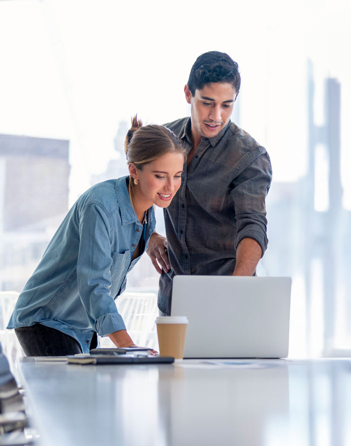 Zwei Personen stehen an einem Tisch und schauen gemeinsam auf einen Laptop. Eine Person zeigt auf den Bildschirm, w&auml;hrend die andere sich interessiert vorbeugt. Die Szene vermittelt Zusammenarbeit und gemeinsames Arbeiten.