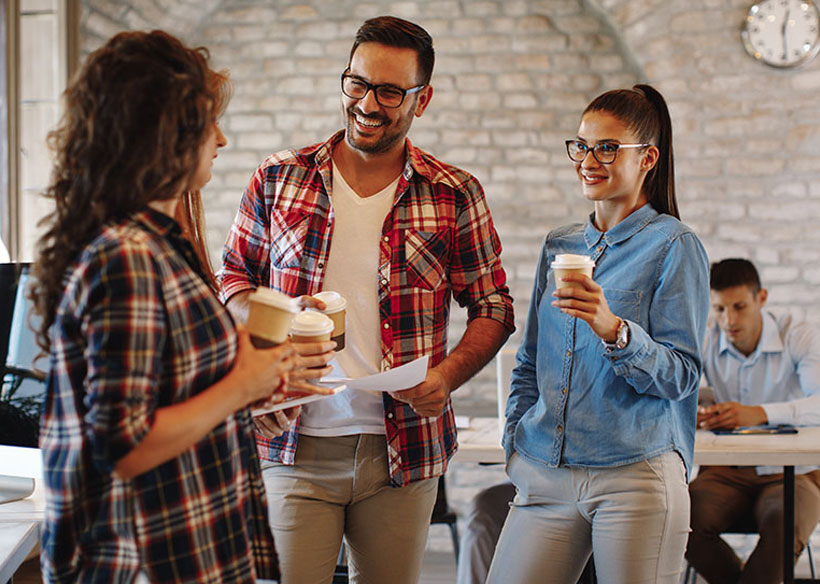 Mehrere Personen stehen in einem B&uuml;ro zusammen, halten Kaffeebecher und unterhalten sich. Eine Person h&auml;lt ein Blatt Papier, die Stimmung wirkt locker und freundlich.