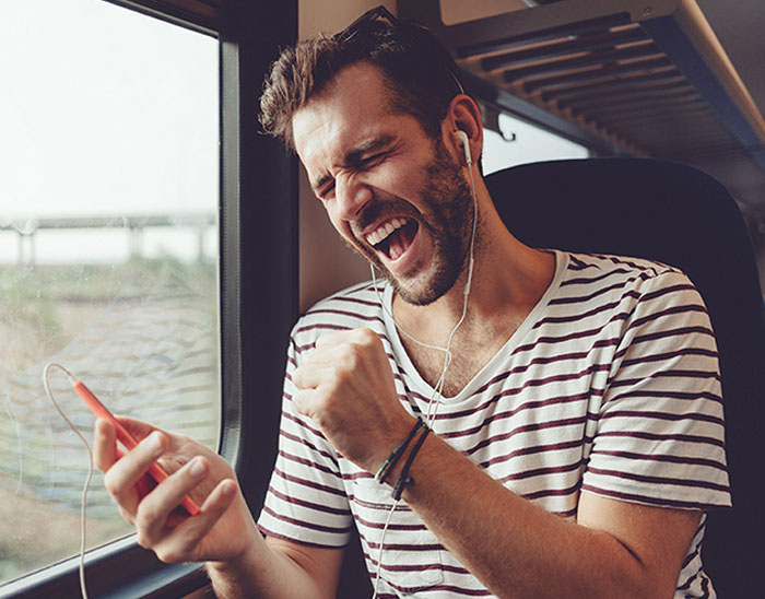 Ein Mann sitzt am Fenster in einem Zug, tr&auml;gt Kopfh&ouml;rer und h&auml;lt ein Smartphone in der Hand. Er wirkt begeistert und singt oder jubelt zur Musik.