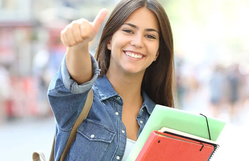 Eine junge Frau h&auml;lt mehrere Ordner im Arm und zeigt mit der anderen Hand den Daumen nach oben. Sie l&auml;chelt und steht im Freien vor einem unscharfen Hintergrund.
