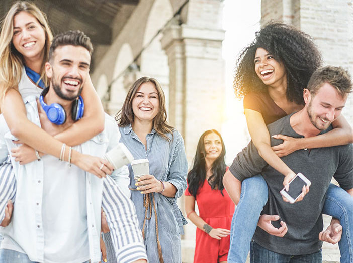 Eine Gruppe junger Menschen geht gemeinsam durch einen &uuml;berdachten Au&szlig;enbereich. Einige lachen, zwei Personen tragen andere huckepack. Die Stimmung wirkt fr&ouml;hlich und gemeinschaftlich.
