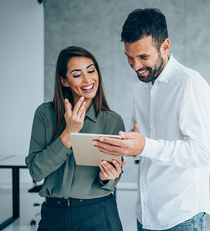 Eine Frau und ein Mann stehen in einem B&uuml;ro und schauen gemeinsam auf ein Tablet. Beide l&auml;cheln und besprechen den angezeigten Inhalt.