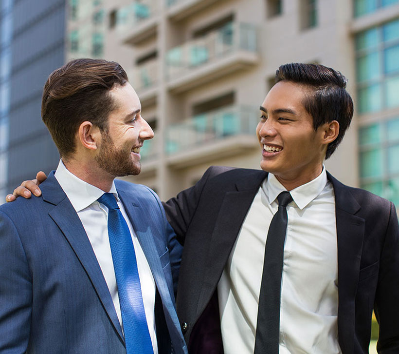 Zwei M&auml;nner im Anzug stehen im Freien vor modernen Geb&auml;uden. Sie legen sich freundschaftlich die Arme um die Schultern und l&auml;cheln sich an.