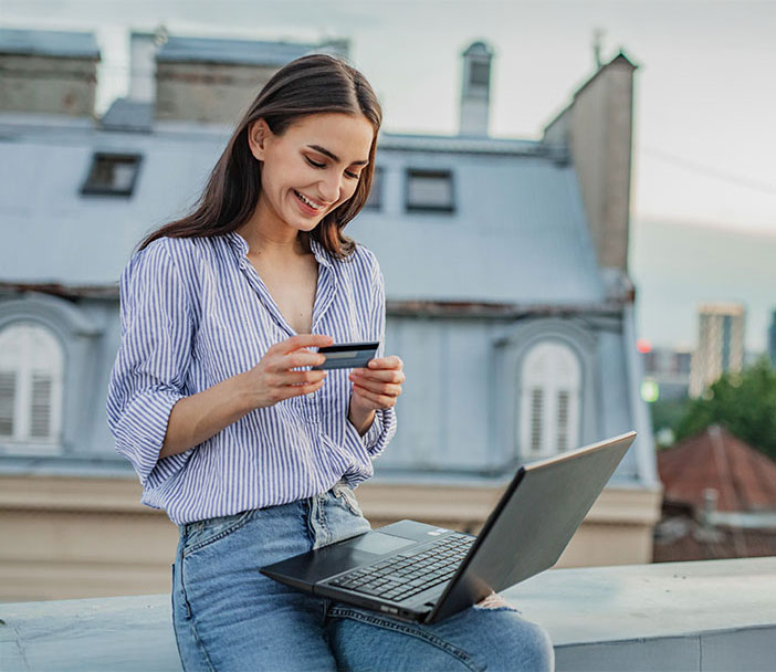 Eine Frau sitzt im Freien mit einem Laptop auf dem Scho&szlig; und h&auml;lt eine Karte in der Hand. Sie l&auml;chelt und schaut auf die Karte, im Hintergrund sind Geb&auml;ude zu sehen.