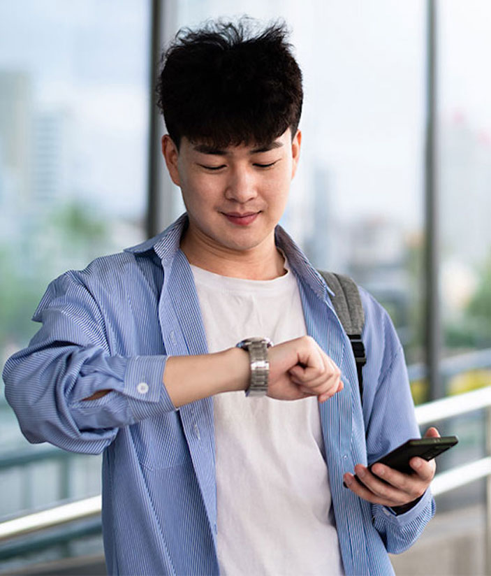 Ein Mann mit Rucksack schaut auf seine Armbanduhr und h&auml;lt ein Smartphone in der Hand. Er steht in einem modernen Umfeld mit Glasfronten im Hintergrund.