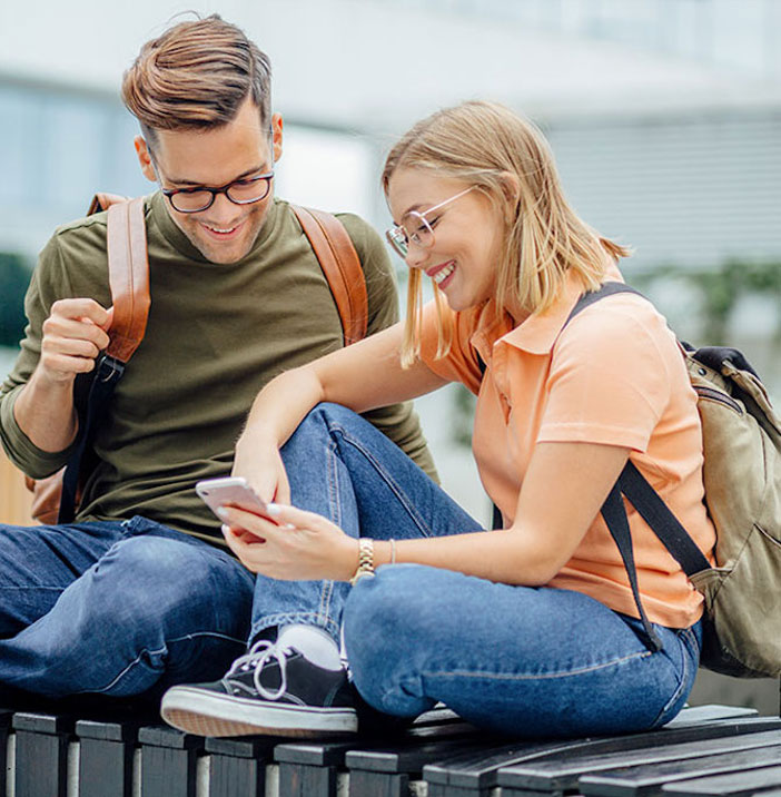 Zwei junge Menschen sitzen nebeneinander auf einer Bank und schauen gemeinsam auf ein Smartphone. Beide tragen Rucks&auml;cke und wirken entspannt und fr&ouml;hlich.